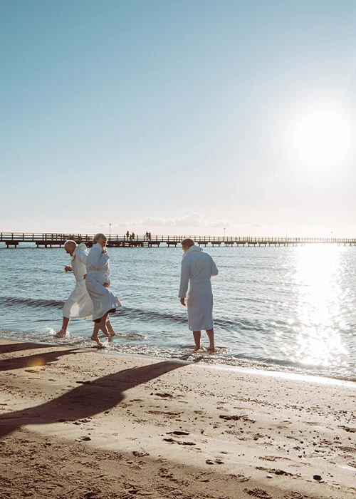 Spring Fling – FSB- Hav Tre personer i vita badrockar springer glatt längs stranden vid Falkenberg Strandbad i solnedgången